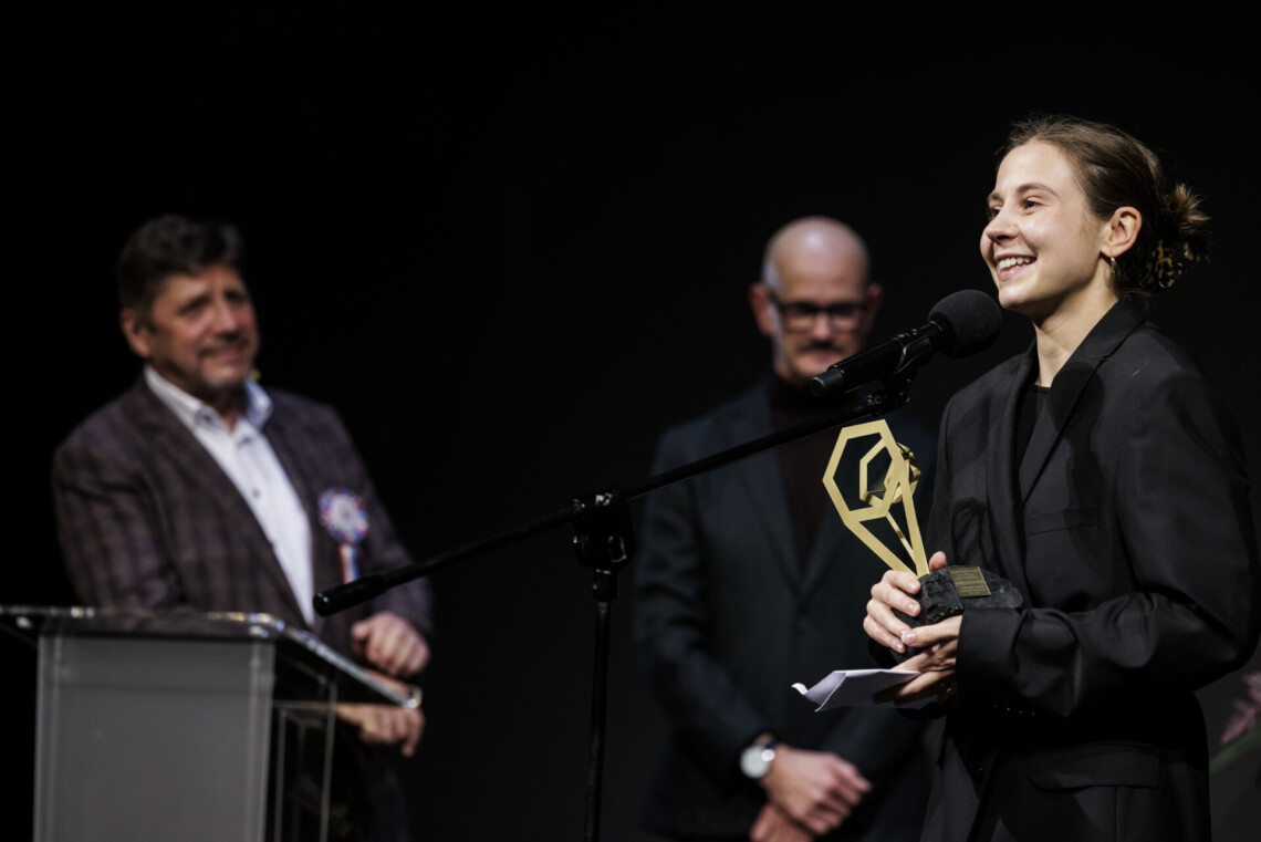 Aleksandra Bernatek przemawia do mikrofonu stojącego na statywie, w dłoniach trzyma nagrodę. W tle rozmyte postaci Dyrektorów Roberta Talarczyka i Bogusława Jasiok.