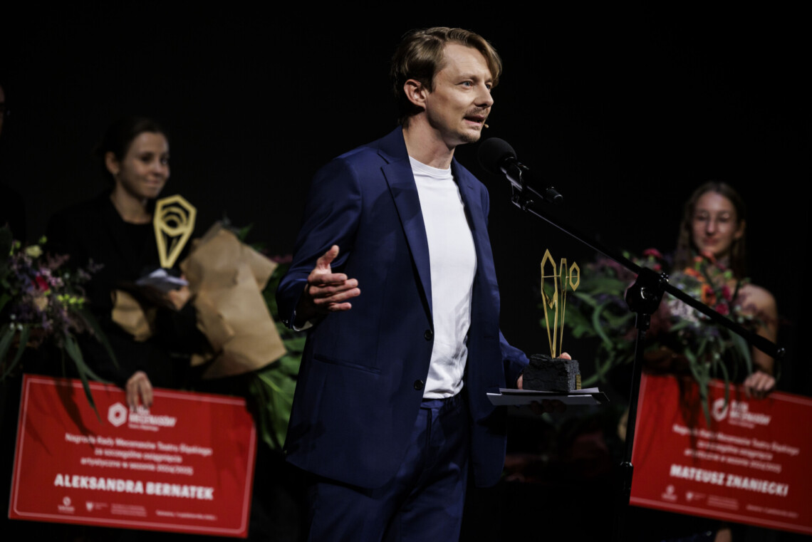 Mateusz Znaniecki przemawia do mikrofonu stojącego na statywie, w lewej dłoni trzyma nagrodę. W tyle stoją Aleksandra Bernatek z nagrodą oraz bileterka z kwiatami.