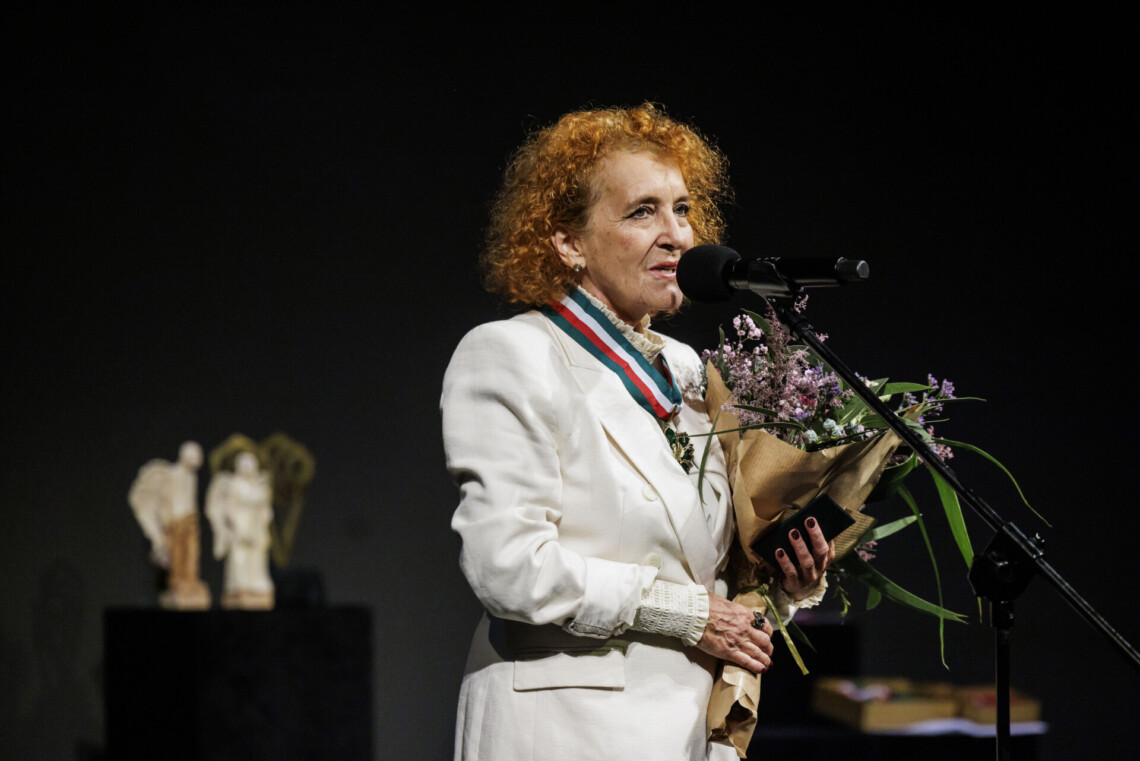 Ewa Leśniak przemawia do mikrofonu stojącego na statywie. Na szyi zawieszony medal, w dłoniach trzyma kwiaty. W tle na kubiku stoją statuetki Angelus Kulis.