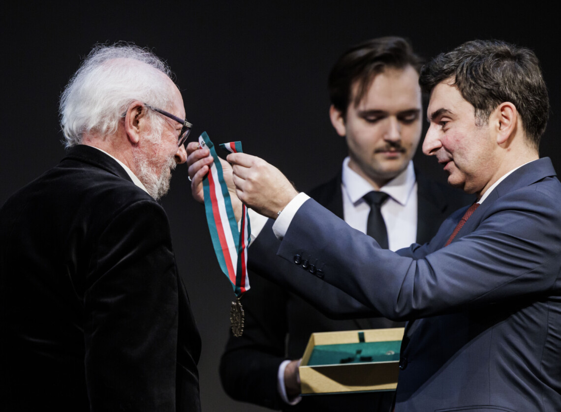 Wojewoda Marek Wójcik zawiesza złoty medal Zasłużony Kulturze Gloria Artis na szyi Romana Michalskiego. W tle rozmyta postać biletera.