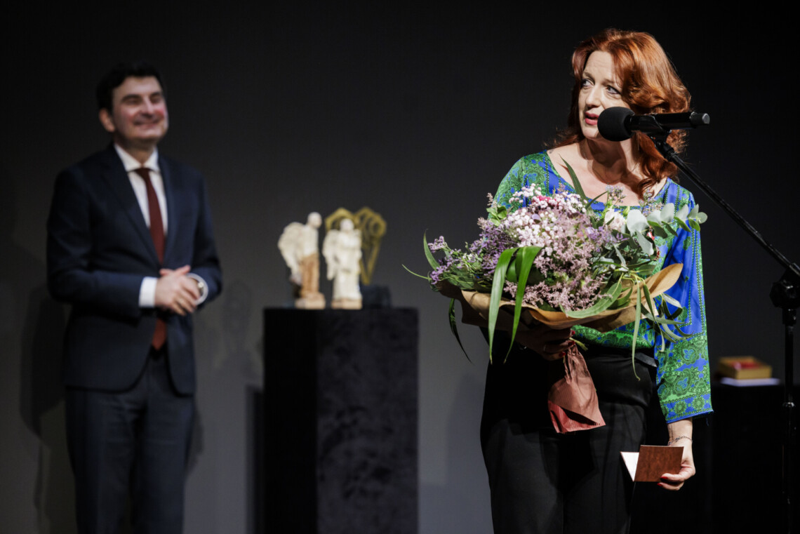 Barbara Lubos przemawia do mikrofonu stojącego na statywie, w prawej dłoni trzyma bukiet kwiatów. Za nią rozmyta postać Wojewody Marka Wójcik oraz statuetki Angelus Kulis stojące na kubikach.