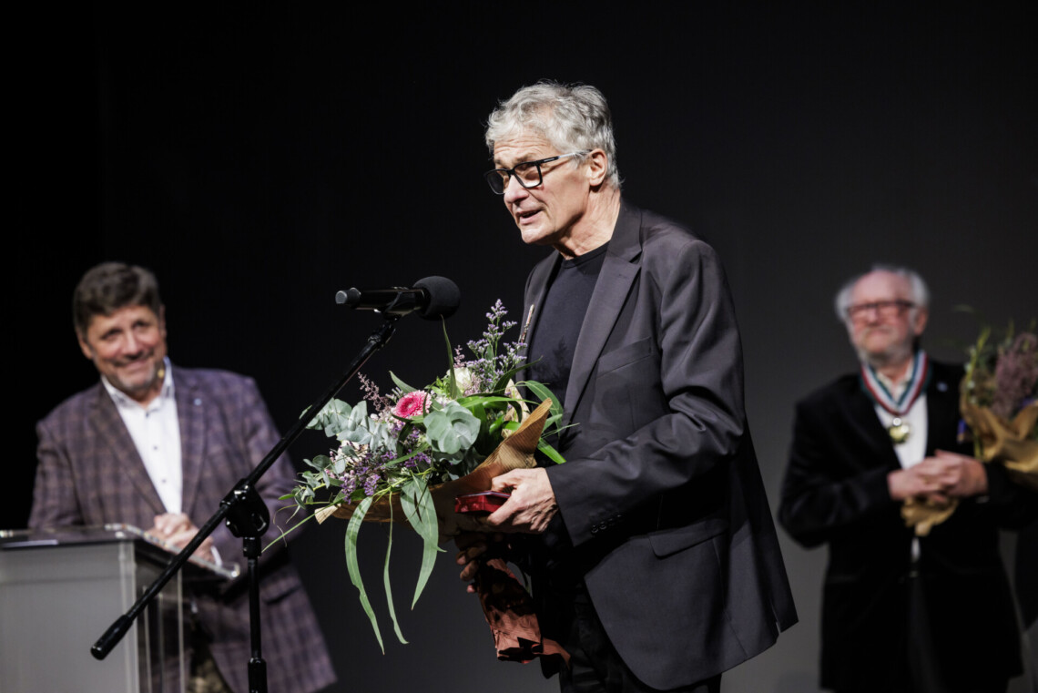 Andrzej Dopierała przemawia do mikrofonu stojącego na statywie. W dłoniach trzyma bukiet kwiatów. W tyle stoi Dyrektor Robert Talarczyk oparty o mównicę oraz rozmyta postać Romana Michalskiego.