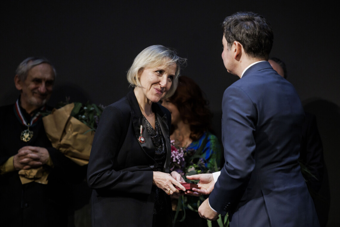 Wojewoda Marek Wójcik Przekazuje Dagmarze Gumowskiej odznaczenie. Za nimi stoją pozostałe odznaczone osoby.