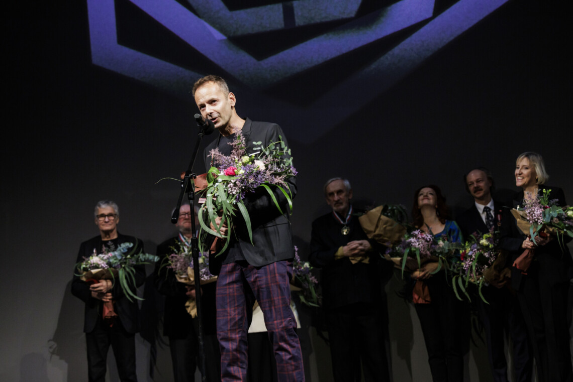 Marcin Szaforz przemawia do mikrofonu stojącego na statywie, w dłoniach trzyma kwiaty. W tle pozostałe nagrodzone osoby.