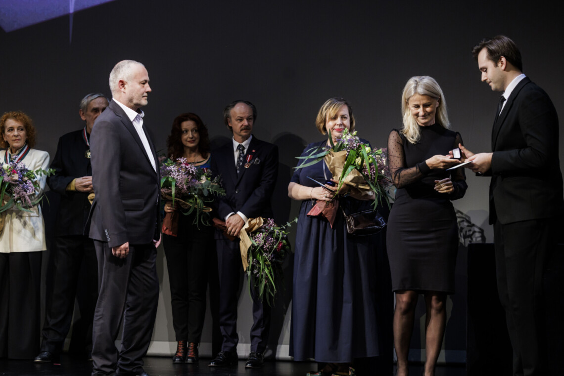 Po lewej Mikołaj Lichtański stoi bokiem do widowni, po prawej Radna Sejmiku Województwa Śląskiego Aleksandra Poloczek odbiera z dłoni biletera odznakę honorową za zasługi dla województwa śląskiego. W tyle stoją pozostałe nagrodzone osoby z bukietami kwiatów w dłoniach.