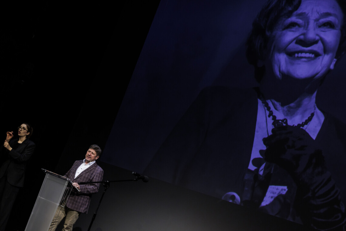 Dyrektor Robert Talarczyk stoi przy mównicy, po jego prawej stronie tłumaczka języka migowego w trakcie pracy. W tle wyświetla się postać Łucji Ginko.