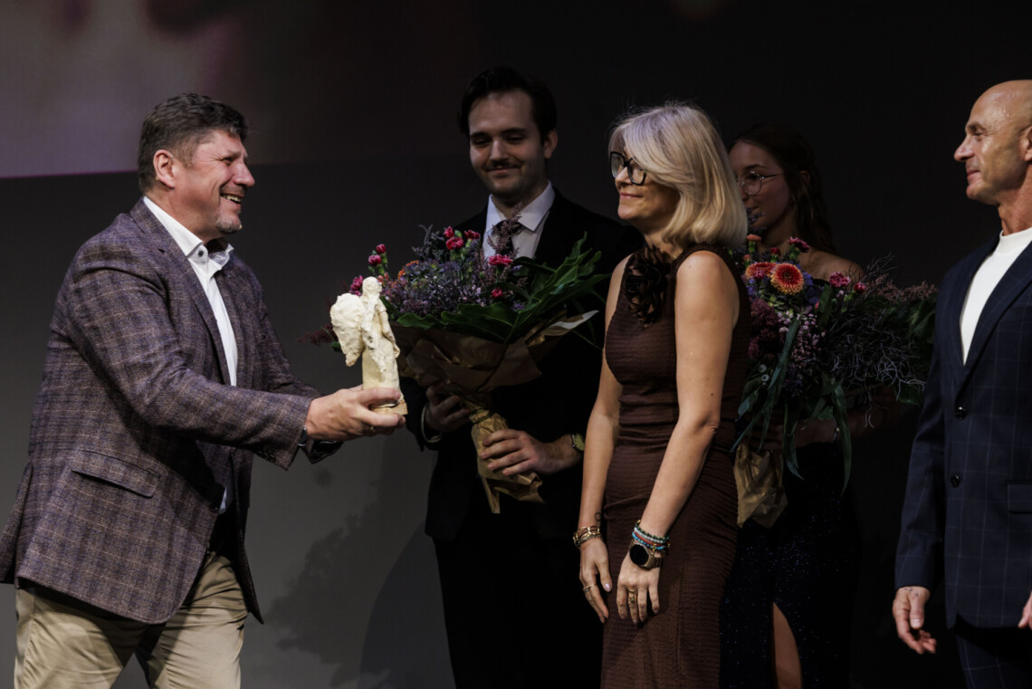 Dyrektor Robert Talarczyk wręcza Wioletcie Krysiak statuetkę Angelius Kulis, obok niej stoi Sebastian Krysiak. W tle para bileterów trzyma bukiety kwiatów.