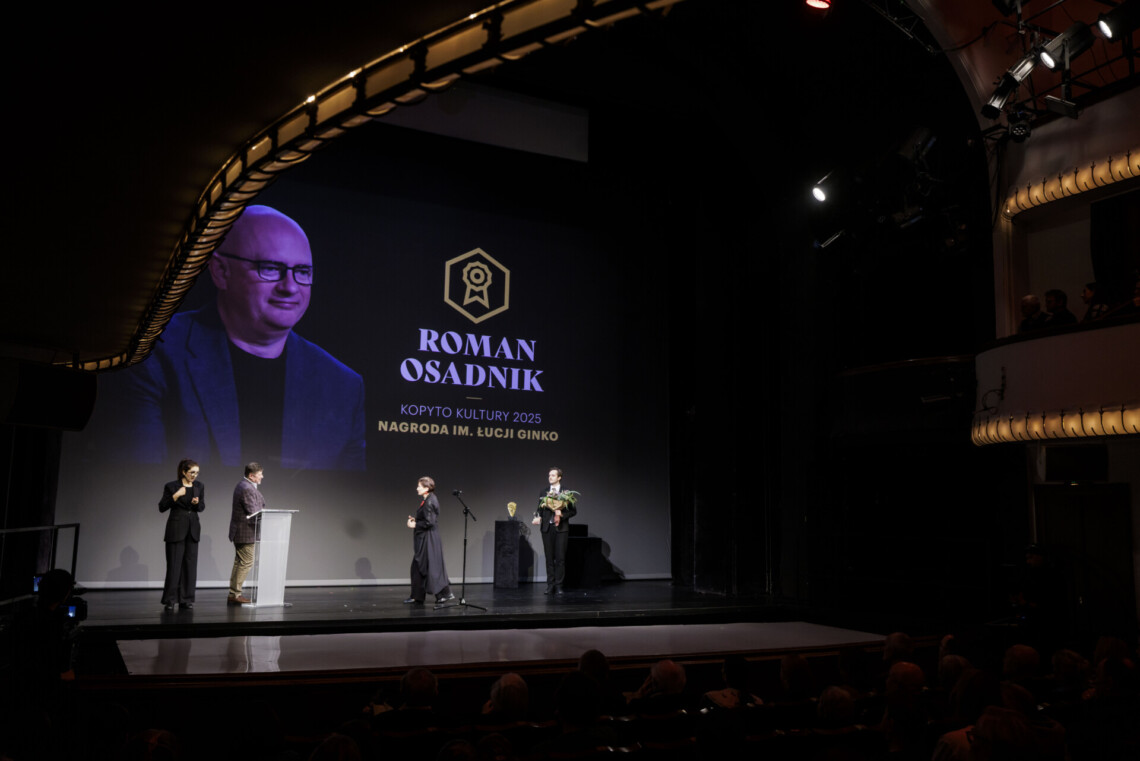 Dyrektor Robert Talarczyk stoi bokiem do mównicy, po jego prawej stronie stoi tłumaczka języka migowego w trakcie pracy. Po lewej kobieta i mężczyzna. W tle wyświetla się postać Romana Osadnika.