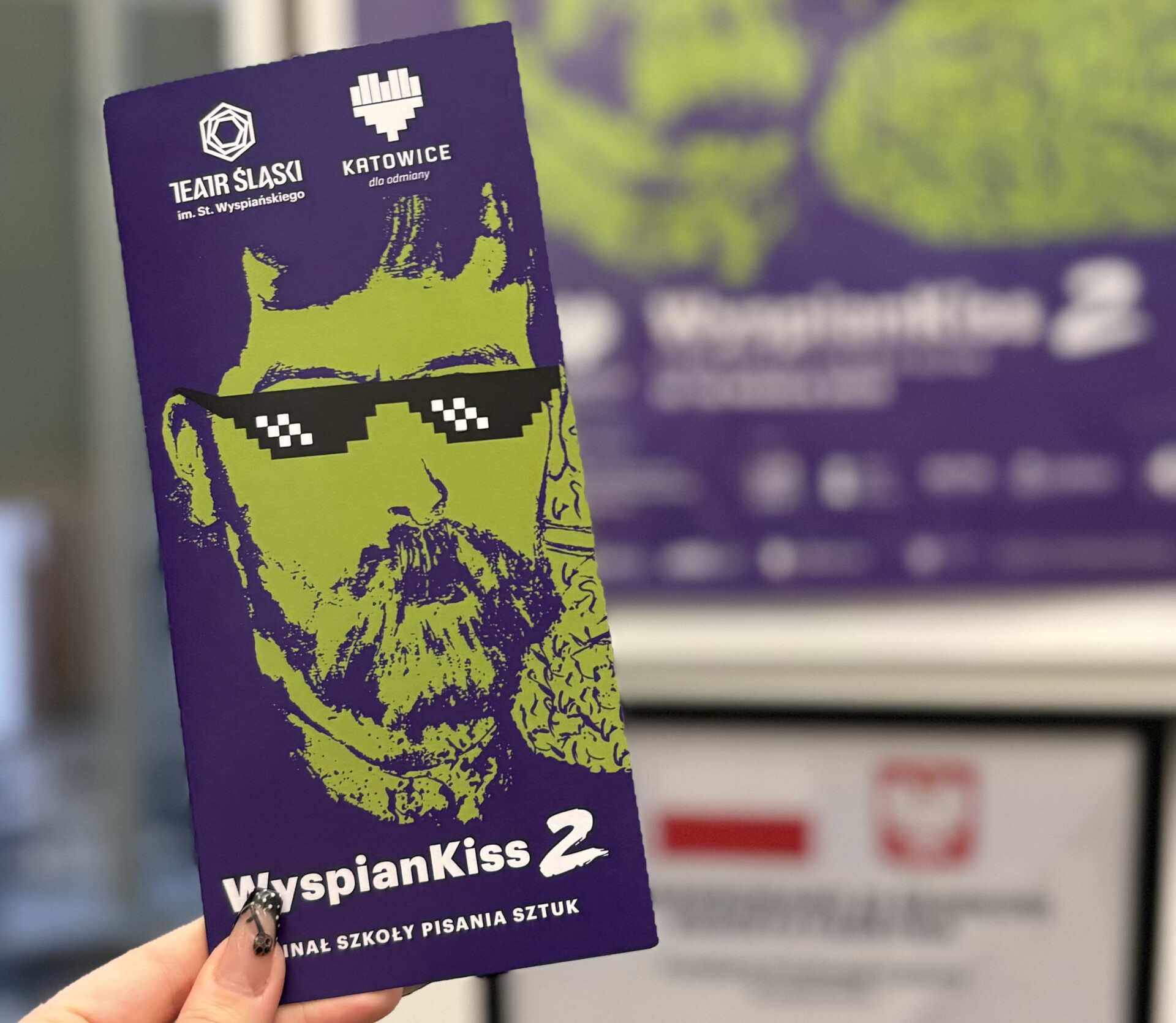 Damska dłoń trzymająca fioletowo-zieloną ulotkę przeglądu WyspianKiss 2. Finał szkoły pisania sztuk.