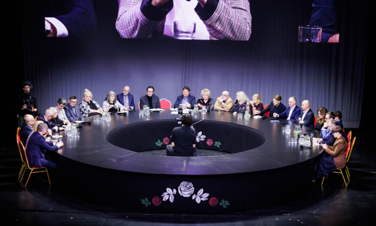 Osoby siedzące wokół czarnego, okrągłego stołu. W środku znajduje się wolna przestrzeń z kamerą i operatorem.