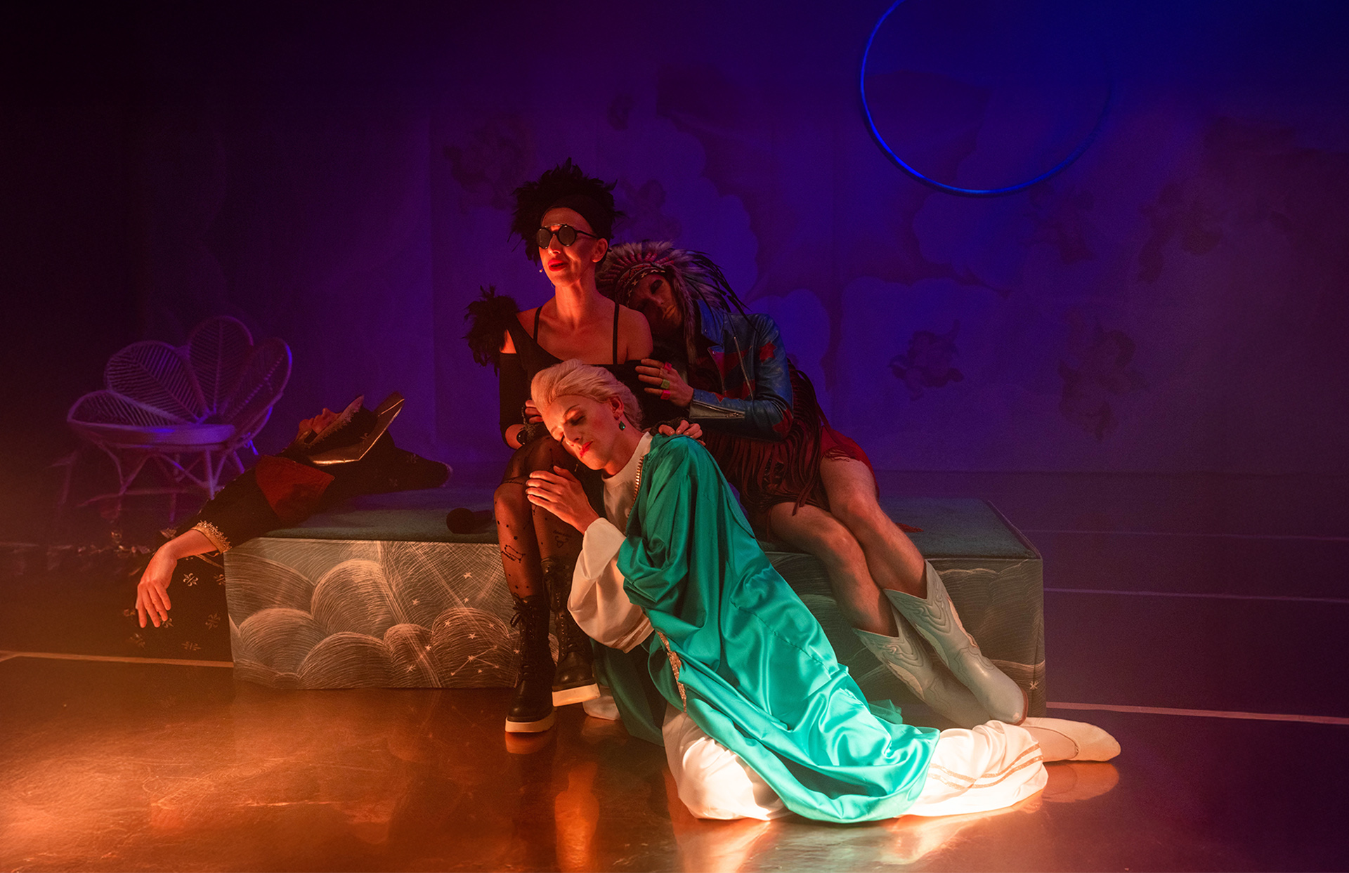 Na podwyższeniu siedzą dwie osoby, trzecia siedzi na podłodze przed nimi i opiera się o nogi jednej z nich, czwarta osoba opiera się plecami o podwyższenie z lewej strony