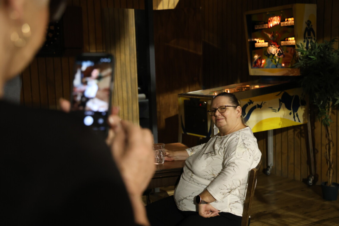 Z lewej strony rozmazana postać osoby robiącej zdjęcie kobiecie siedzące przy drewnianym stoliku i trzymającej w dłoni kufel, w tle automat do gry zręcznościowej, osoby znajdują się w barze stanowiącym scenografię spektaklu Tkocze