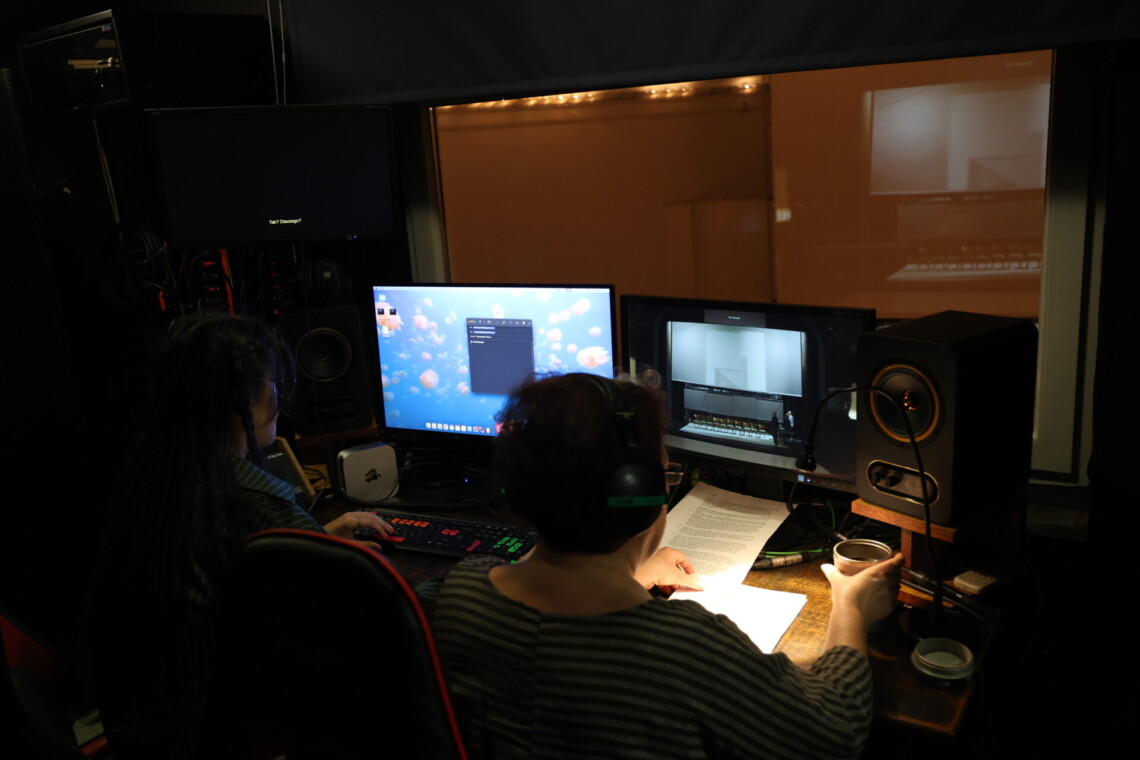 Dwie osoby siedzące przed dwoma monitorami, na prawym widoczny podgląd sceny ze scenografią spektaklu Tkocze, ujęcie od tyłu