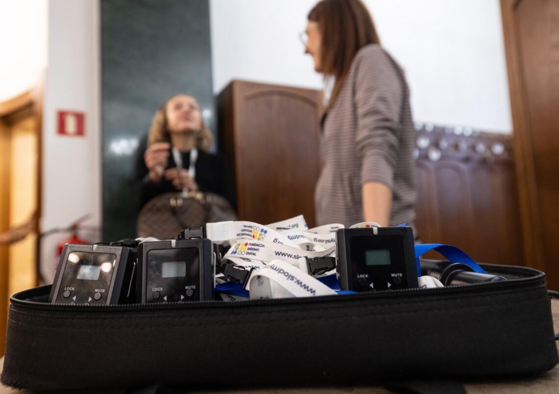 Odbiorniki do audiodeskrypcji leżące w futerale na ladzie, w tle rozmazane postacie dwóch osób