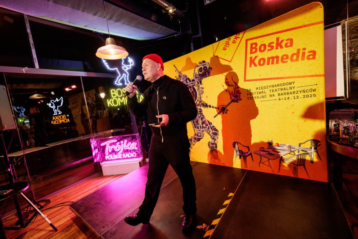 Bartosz Szydłowski Dyrektor Artystyczny Festiwalu Boska Komedia przemawia do mikrofonu na tle ścianki reklamowej Festiwalu w kolorze żółtym