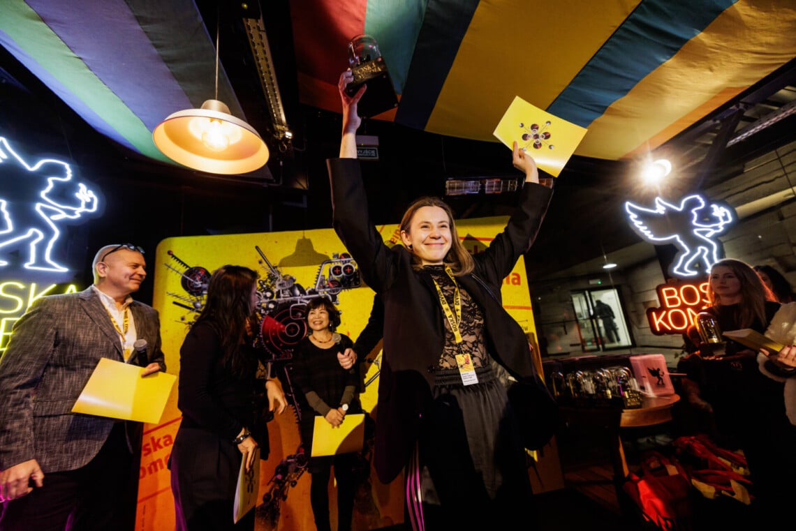 Aleksandra Bernatek trzyma w prawej dłoni nagrodę, w lewej żółty skoroszyt, unosi je wysoko nad głową