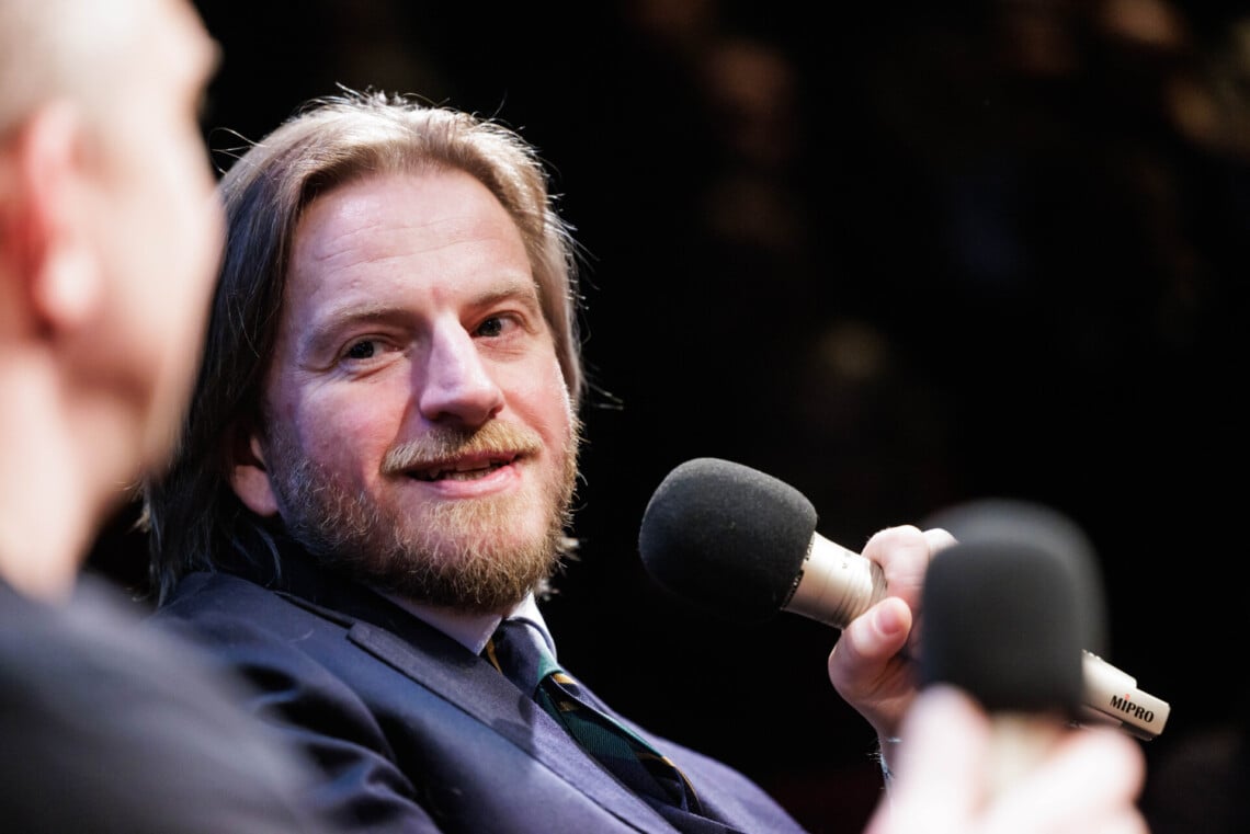 Szczepan Twardoch trzyma w dłoni mikrofon, patrzy w bok w kierunku swojego rozmówcy, którego widać tylko niewyraźny profil, ujęcie z bliska