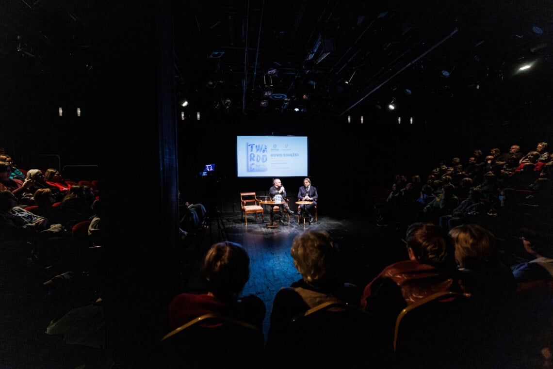 W centrum dwa okrągłe stoliki i trzy krzesła, na dwóch siedzą mężczyźni, za nimi ekran z wyświetlonym plakatem promującym spotkanie, widzowie siedzą z prawej, lewej i z przodu sceny