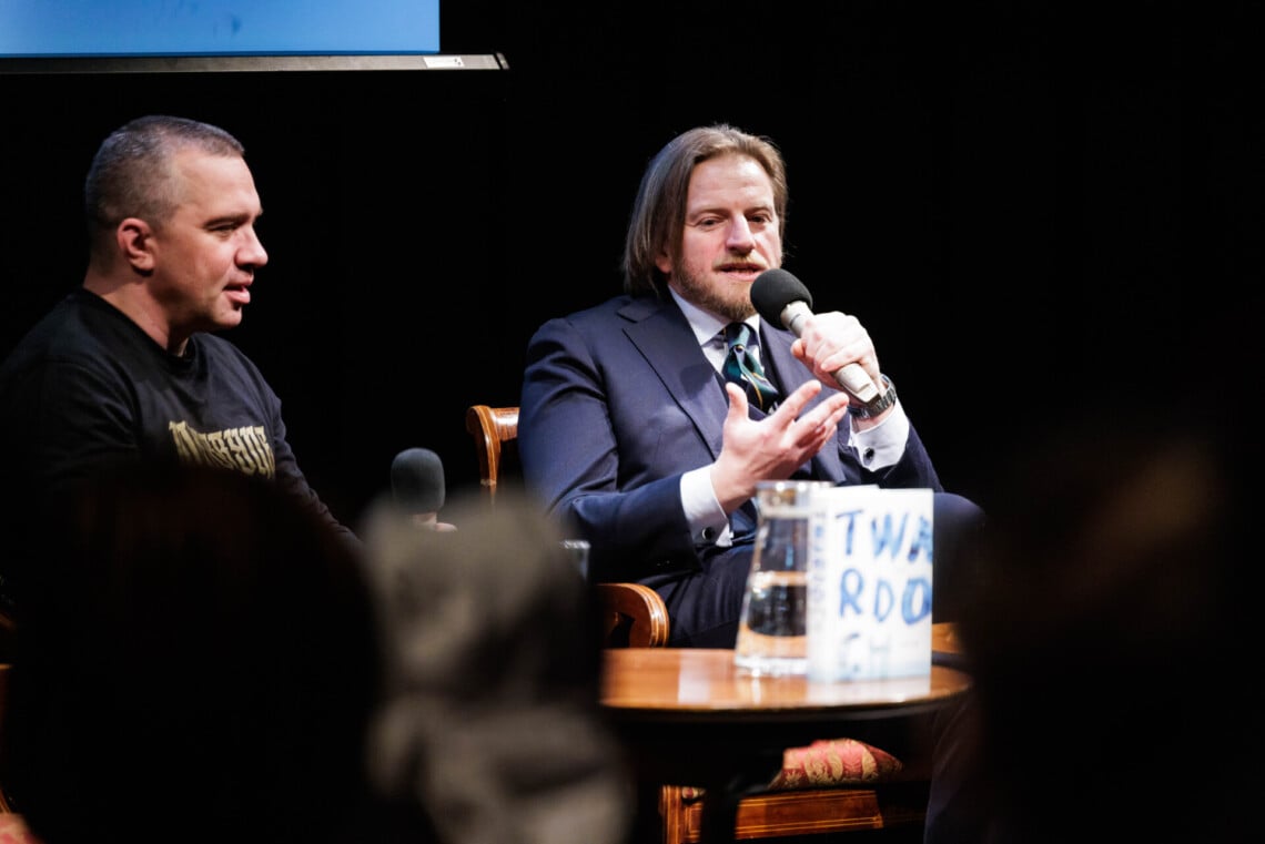 Łukasz Orbitowski oraz Szczepan Twardoch siedzą na krzesłach, do mikrofonu przemawia gość