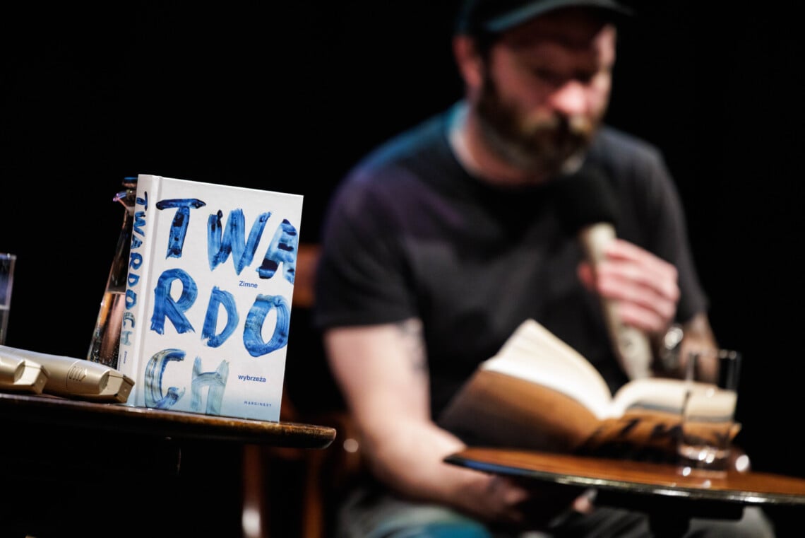Na stoliku postawiono pionowo książka, na okładce słowo TWARDOCH drukowanymi niebieskimi literami jakby malowanymi pędzlem, w tle rozmazana postać mężczyzny z książką i mikrofonem