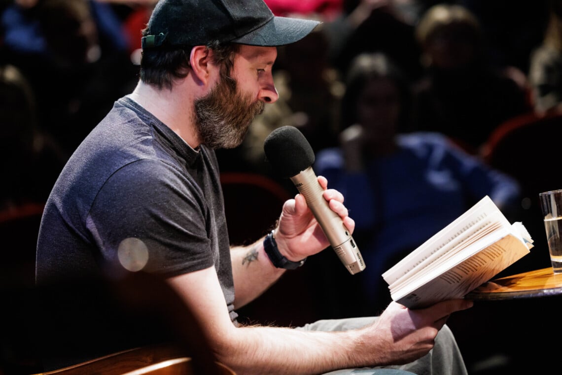 Aktor Dariusz Chojnacki czytający fragment książki, w prawej dłoni trzyma otwartą książkę, w lewej mikrofon, ujęcie z boku