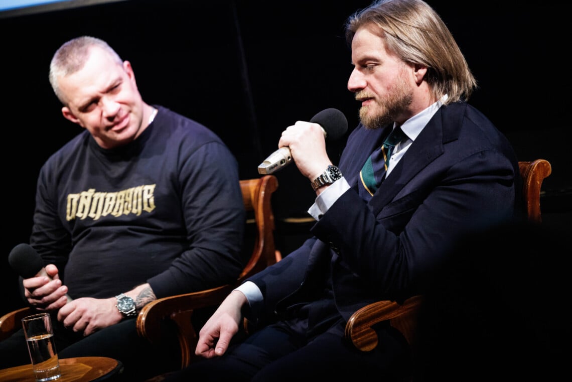 Szczepan Twardoch oraz Łukasz Orbitowski siedzą na krzesłach, do mikrofonu przemawia gość, ujęcie z boku