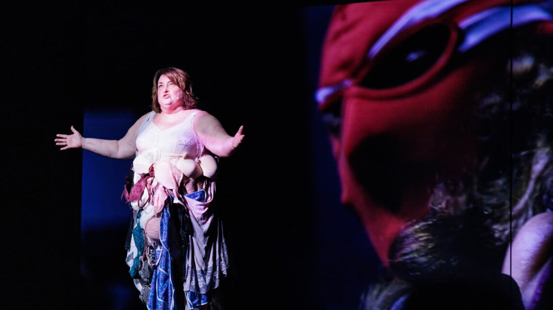 Na scenie kobieta w biustonoszu obwieszona różnymi biustonoszami i podomkami, które tworzą spódnicę, stoi z lewej strony, z prawej w tle fragment ekranu na którym widać twarz w czerwonej masce