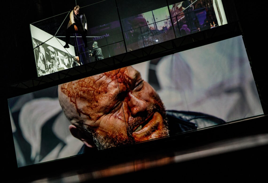 Na dole na podłużnym ekranie wyświetlona twarz zakrwawionego mężczyzny z przymkniętymi oczami, powyżej na piętrze scena, przed nią w powietrzy znajduje się zawieszony człowiek