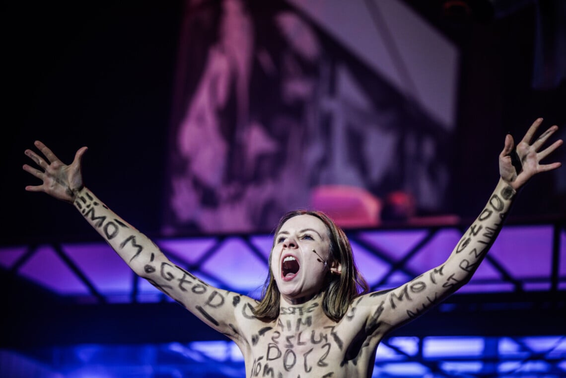 Kobieta bez koszulki z zapisanymi na ciele czarnymi napisami "mam głód", ma uniesione w górę ręce i szeroko otwarte usta, zdjęcie powyżej klatki piersiowej