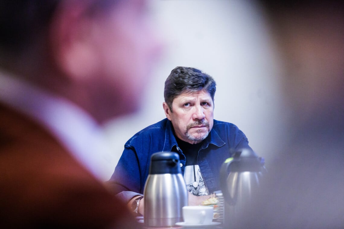 Dyrektor Robert Talarczyk siedzący za stołem, ujęcie z daleka, za pierwszym planie rozmazana postać mężczyzn, dyrektor jest jakby między nimi