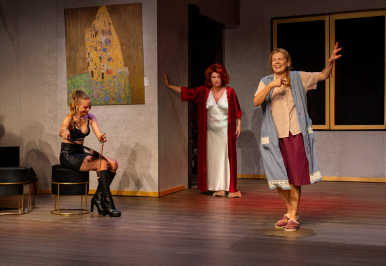 Trzy kobiety w pomieszczeniu, pierwsza ubrana na czarno siedzi na czarnej pufie z lewej, z prawej stoi kobieta w fartuszku, po środku z tyłu opierając się o ścianę stoi czwarta w eleganckiej koszuli nocnej i w szlafroku