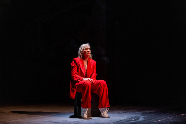 Na betonowym kamieniu siedzi pięćdziesięcioparoletnia kobieta. Ubrana jest elegancko, w czerwony garnitur. Jej usta są pomalowane na ten sam kolor, są rozchylone, mówi coś. Ręce opiera na kolanach. W jednej z nich trzyma papierosa, z którego unosi się dym obok jej głowy. Tło czarne.